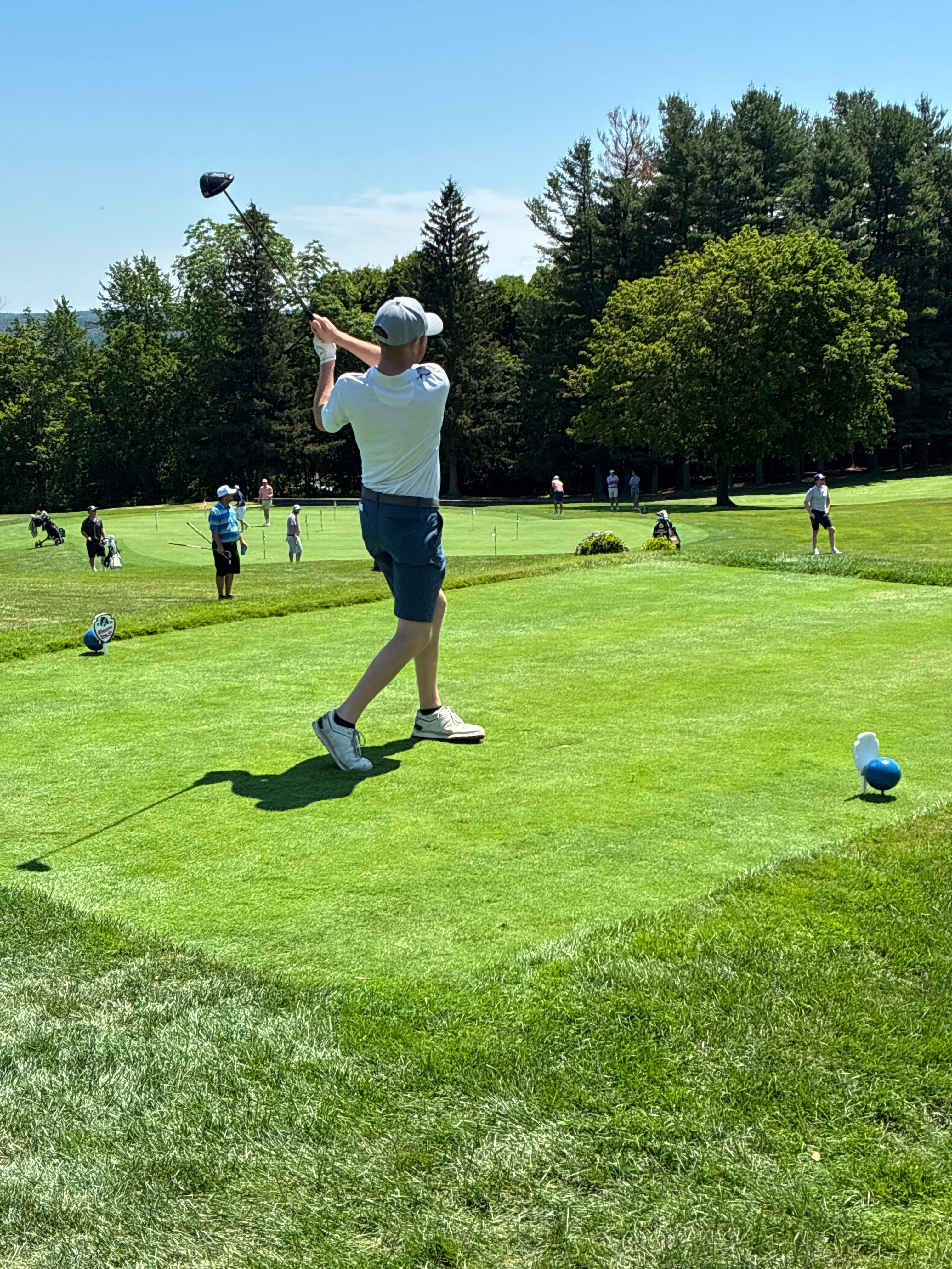 Professional golfer mid-swing at golf tournament with custom tee signs visible on course